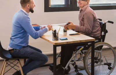AccessiBe Ltd, two person are sitting face to face on table talk. One is disabled and sitting on wheel chair
