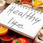 Side view of a healthy life inscription on spiral notebook on fresh vegetables lemon corn kernels lemon fallen oil bottle honey on white background