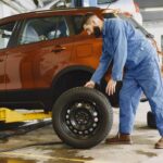 Man in Blue Coveralls Standing Beside an Orange Car Checking on the Tire