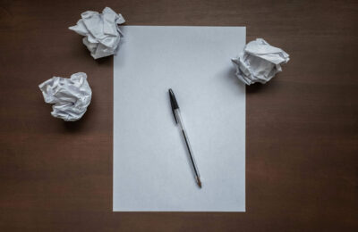 Flat lay with blank writing sheet, pen and crumpled paper balls on top of wooden office desk. Writer's block or creativity crisis concept. Top down view