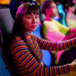 A Woman in Striped Long Sleeves Sitting on a Car Arcade Machine while Seriously Looking at the Camera