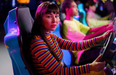A Woman in Striped Long Sleeves Sitting on a Car Arcade Machine while Seriously Looking at the Camera
